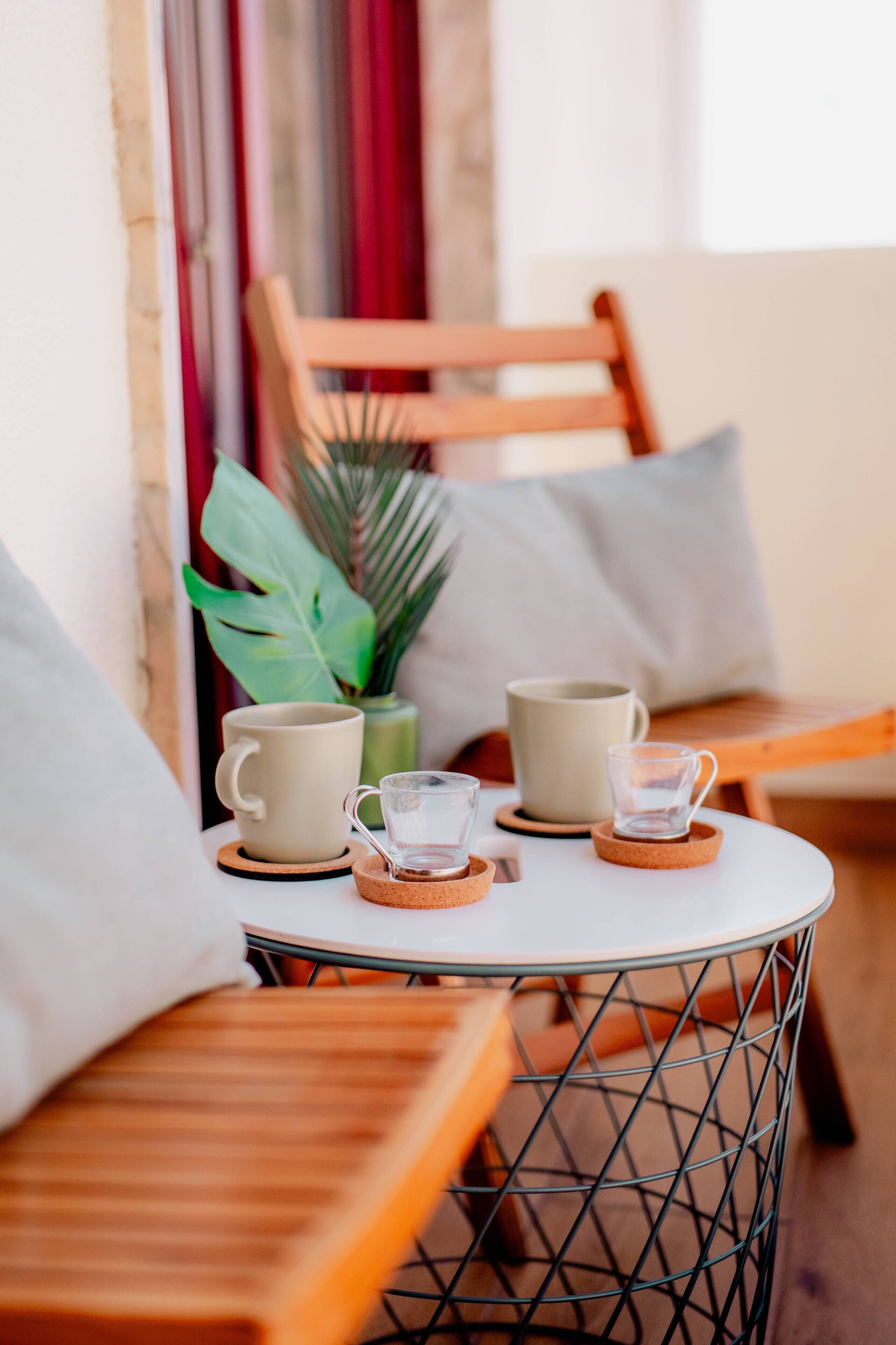 A cozy seating area with a round metal table, two beige mugs, and a glass cup on coasters. A wooden chair and a potted plant are in the background.