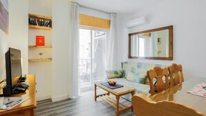 Een woonkamer met een bank, houten stoelen, een glazen salontafel, een televisie en een boekenkast.