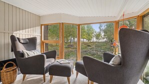 Une pièce chaleureuse avec deux fauteuils gris, un petit pouf et un panier en osier. De grandes fenêtres offrent une vue sur une zone boisée.
