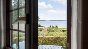A view through a window with a wooden table and chairs, a small wooden structure, and a body of water with a distant shoreline. - Nice apartment in Egernsund with WiFi (Egernsund)