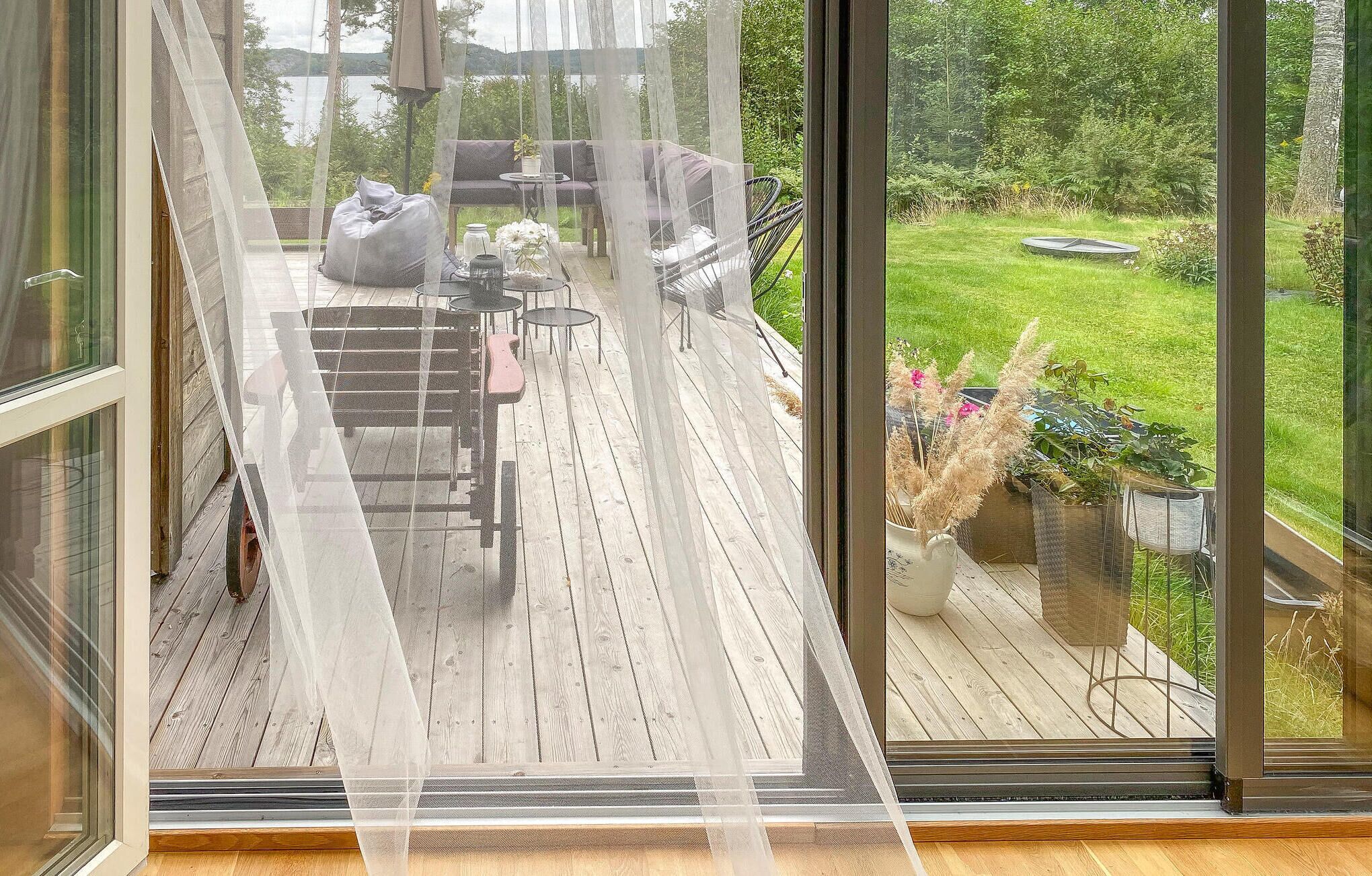 En terrass med trägolv, en bänk, ett bord och stolar. Utsikten sträcker sig över en gräsyta med träd och en vattenyta i bakgrunden.