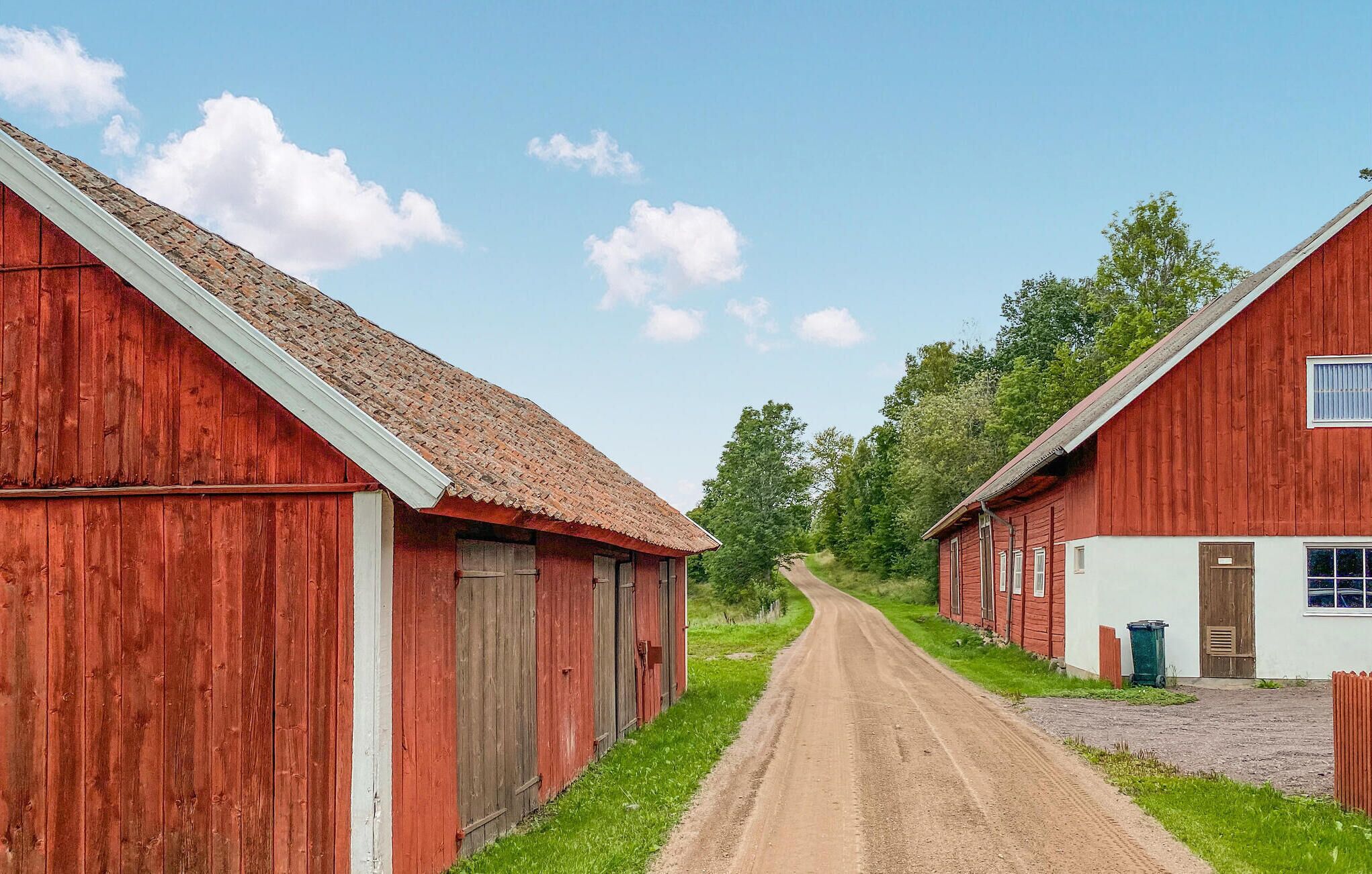 Un camino rural bordeado por edificios de madera roja con detalles blancos, un sendero de tierra y árboles verdes al fondo.