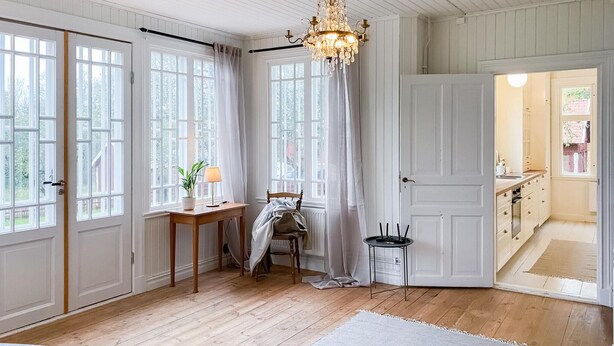 A room with large windows, a chandelier, a desk with a lamp, a chair, and a door leading to another room.