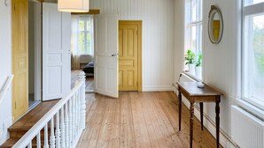 A hallway with wooden flooring, white walls, and a wooden door. There is a white railing on the left, a table with a plant on the right, and a window with a mirror on the wall.