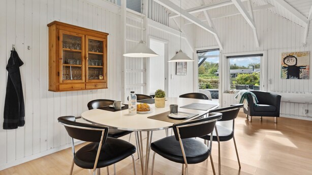 A spacious dining area with a white table and black chairs, a wooden cabinet with glass doors, and a view of greenery outside.
