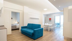 Un salon moderne avec un canapé bleu, un coin repas avec une table et des chaises blanches, et un espace cuisine équipé d’un réfrigérateur.