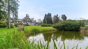 Une grande maison avec un étang au premier plan, entourée d’une végétation luxuriante et d’arbres.