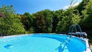 An outdoor swimming pool with a clear blue water surface, surrounded by a metal fence and lush green trees in the background.