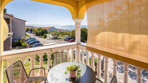 A balcony with a view of parked cars and buildings, a round table with a potted plant, and a white balustrade.