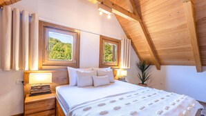 Ein Schlafzimmer mit Holzboden, zwei Fenstern mit Vorhängen, einem Bett mit weißer Bettwäsche, einer Nachttischlampe und einem kleinen Nachttisch.