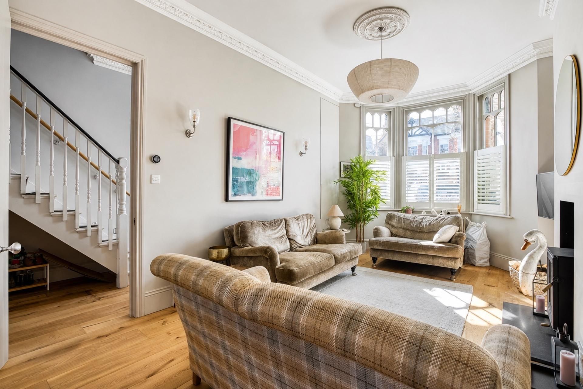 A spacious living room with a staircase, a large window with shutters, and a modern chandelier.