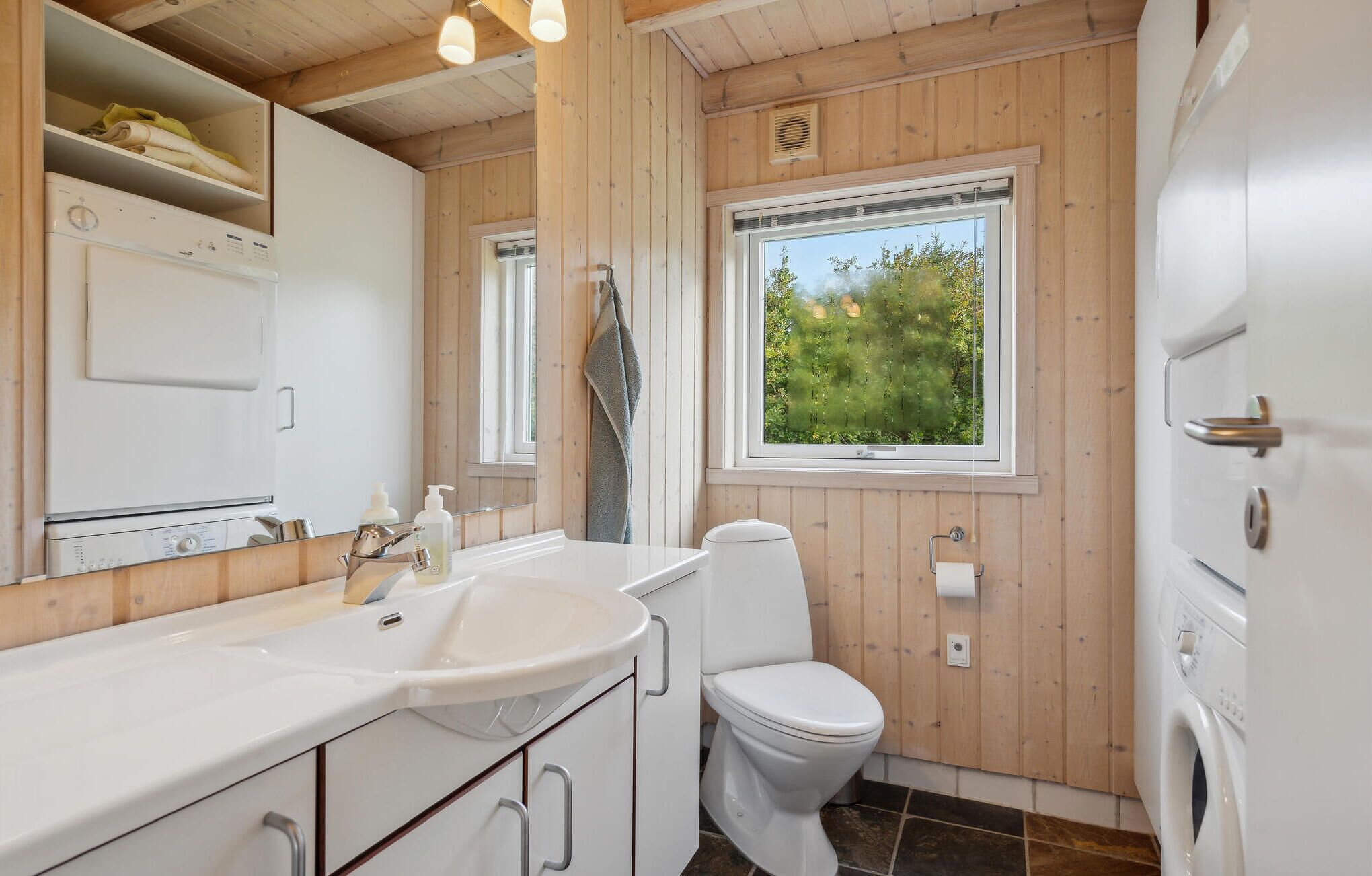 A bathroom with a sink, mirror, toilet, and a window with a view of greenery.