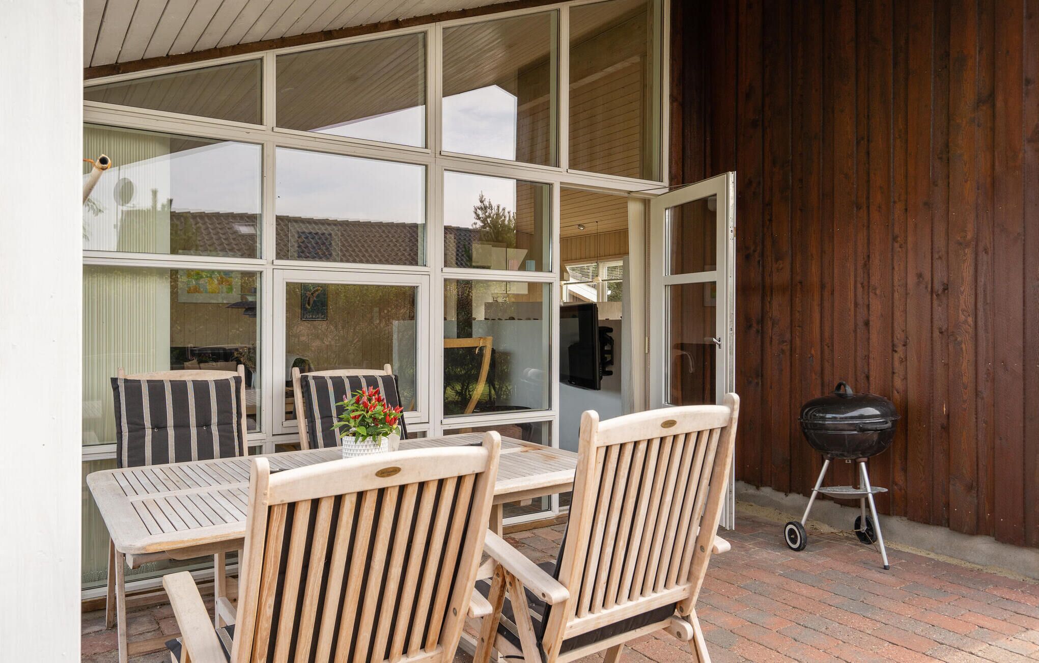 Une terrasse extérieure avec une table et des chaises en bois, un barbecue et une vue sur l’intérieur, visible à travers de grandes fenêtres.
