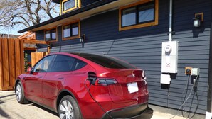 Une Tesla Model 3 rouge est garée devant une maison, qui est entourée d’une clôture en bois et à côté de laquelle se trouve un coffret électrique.