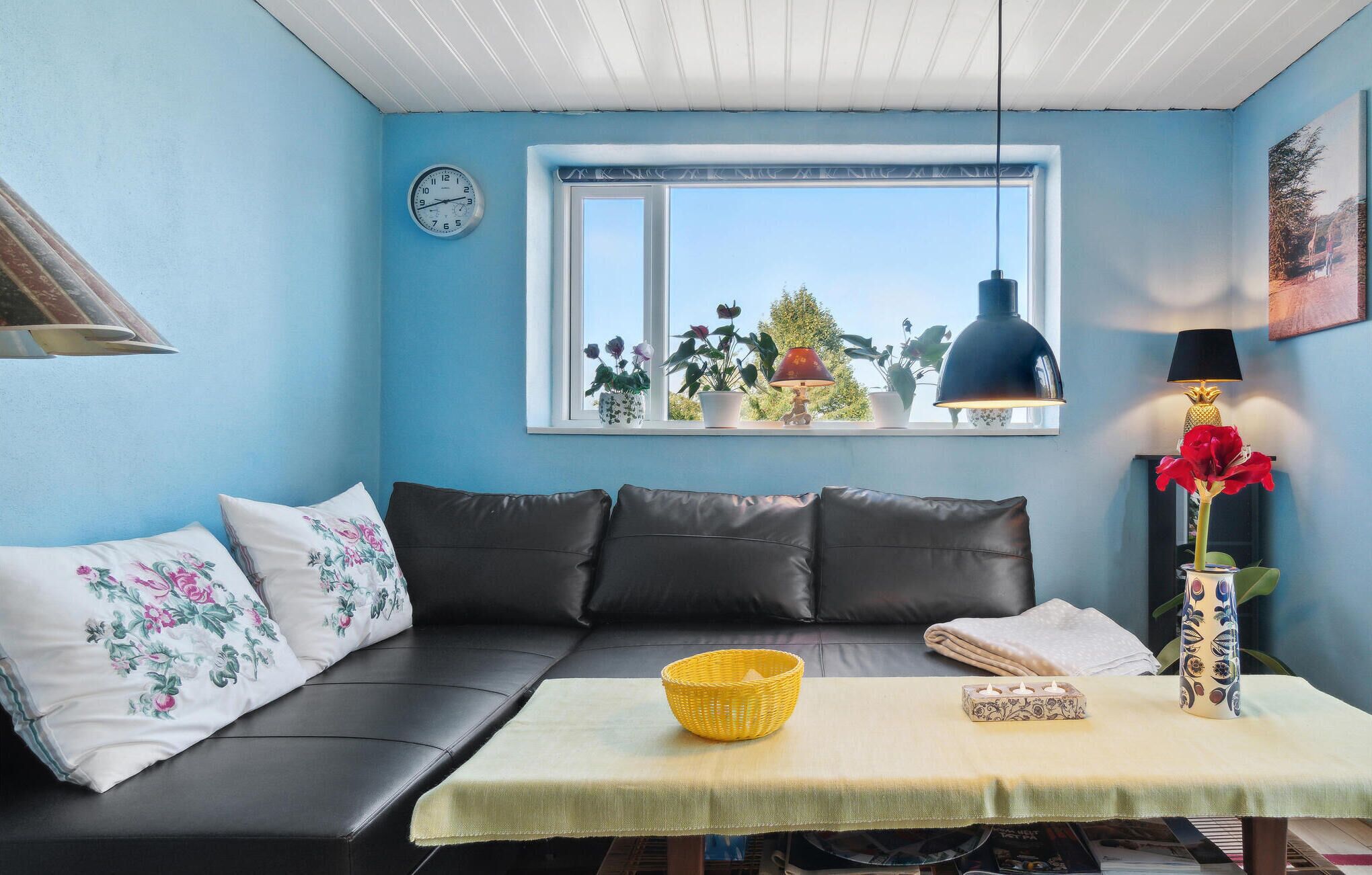 Ett mysigt rum med en blå vägg, en svart lädersoffa, ett litet bord med en gul korg och ett fönster med växter.