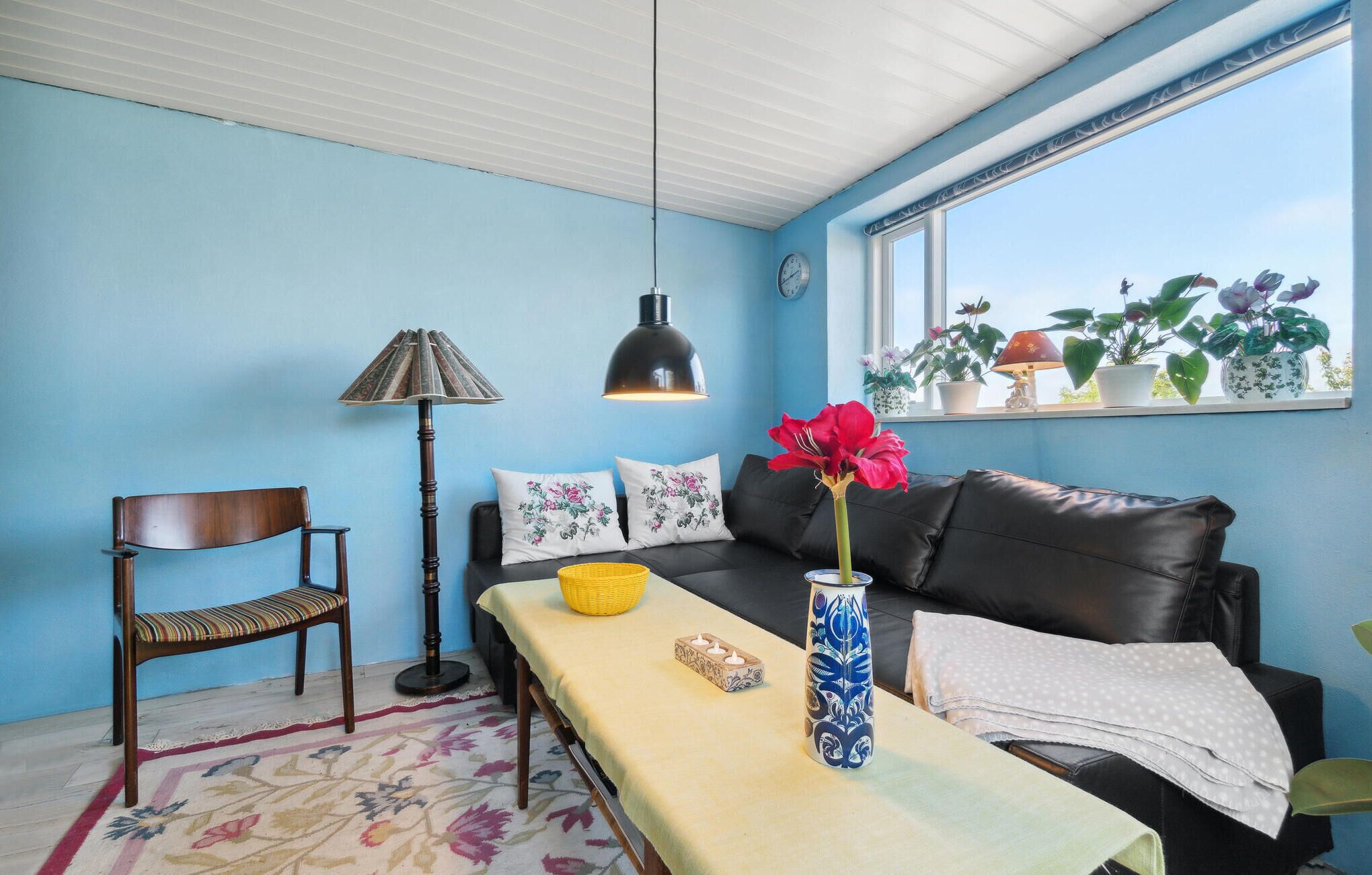A living room with a sofa, a table with a vase, a chair, a lamp, and a window with plants.