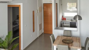 Une chambre d’hôtel compacte comprenant un coin cuisine, un espace repas avec une table et des chaises en bois, une petite salle de bain avec douche, et une porte donnant sur une autre pièce.