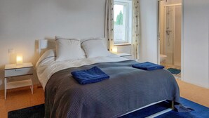 Un lit bien fait, recouvert d’une couverture bleue, une lampe de chevet et une fenêtre donnant sur un paysage verdoyant.