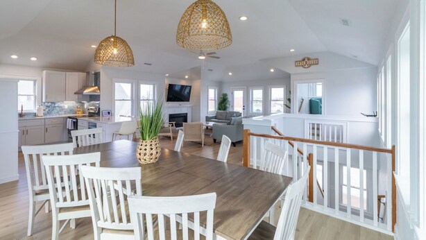 A spacious kitchen with white cabinetry, a dining area with a wooden table and white chairs, and a living area with a sofa and TV. - 🏖️1 min to beach access!🏊‍♂️Heated pool, hot tub, pets, ocean views, arcade! (Nags Head)