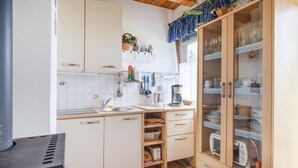 Cocina compacta con armarios blancos, un lavamanos y un tostador. Se observa una estantería con utensilios de cocina y una ventana con cortina.