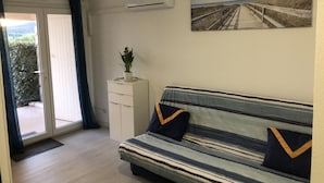 A living room with a striped sofa, a small white cabinet with a plant, and a painting of a beach walkway. - Nouvel Grand Studio Agay vue mar Avec Jardin (Saint-Raphaël)