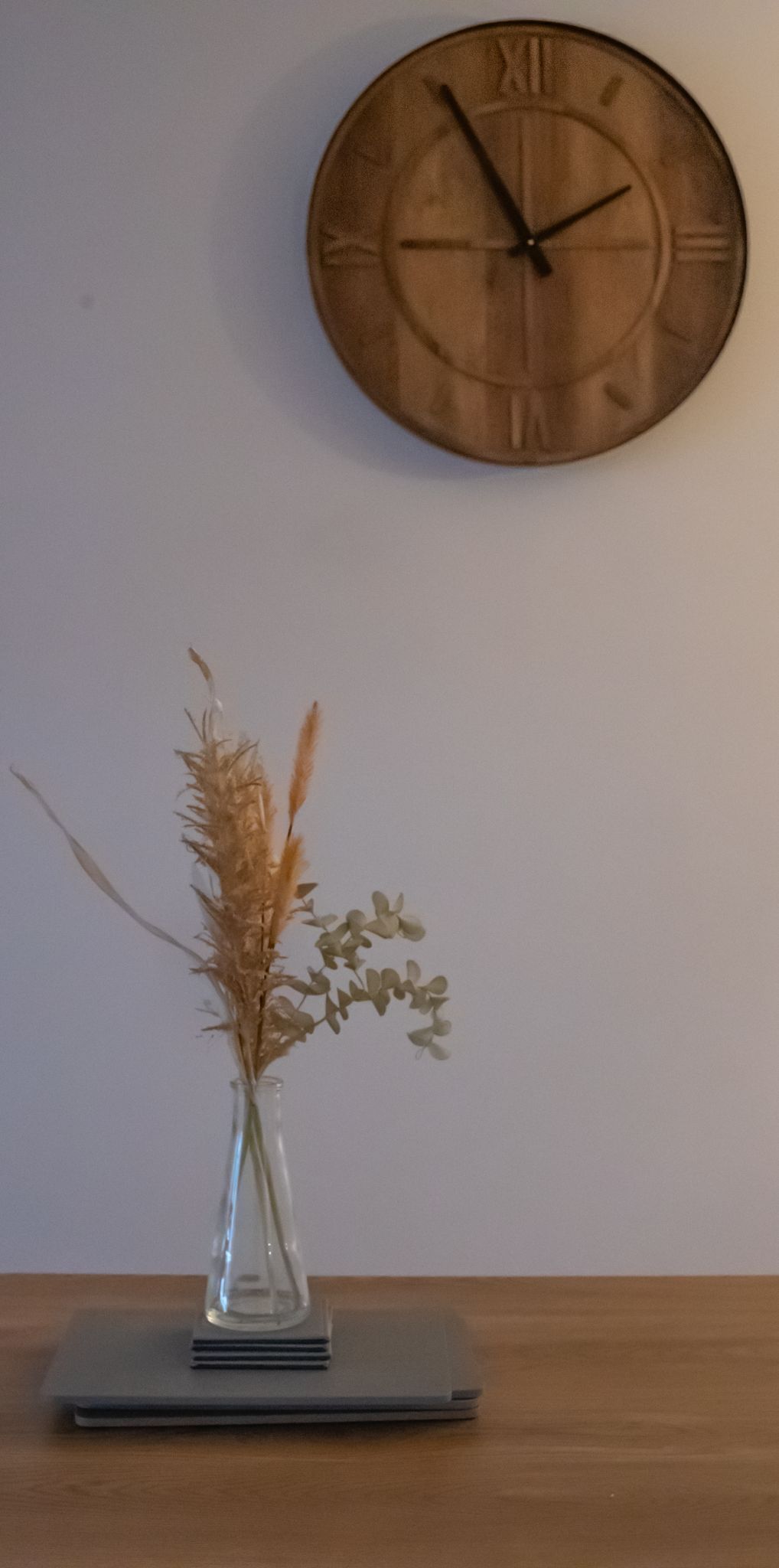 A clock on the wall and a vase with dried flowers on a table.