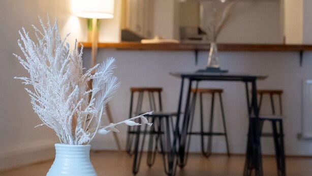 A white vase with dried plants on a wooden table in a room with a kitchen area in the background. - Beautiful 1-bed Apartment in Manchester (Manchester)