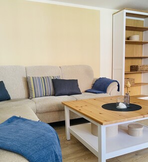 Ein Wohnzimmer mit einem beigen Sofa, einem gestreiften Kissen, einer blauen Decke, einem Holztisch und einem weißen Bücherregal mit Korbwäsche.