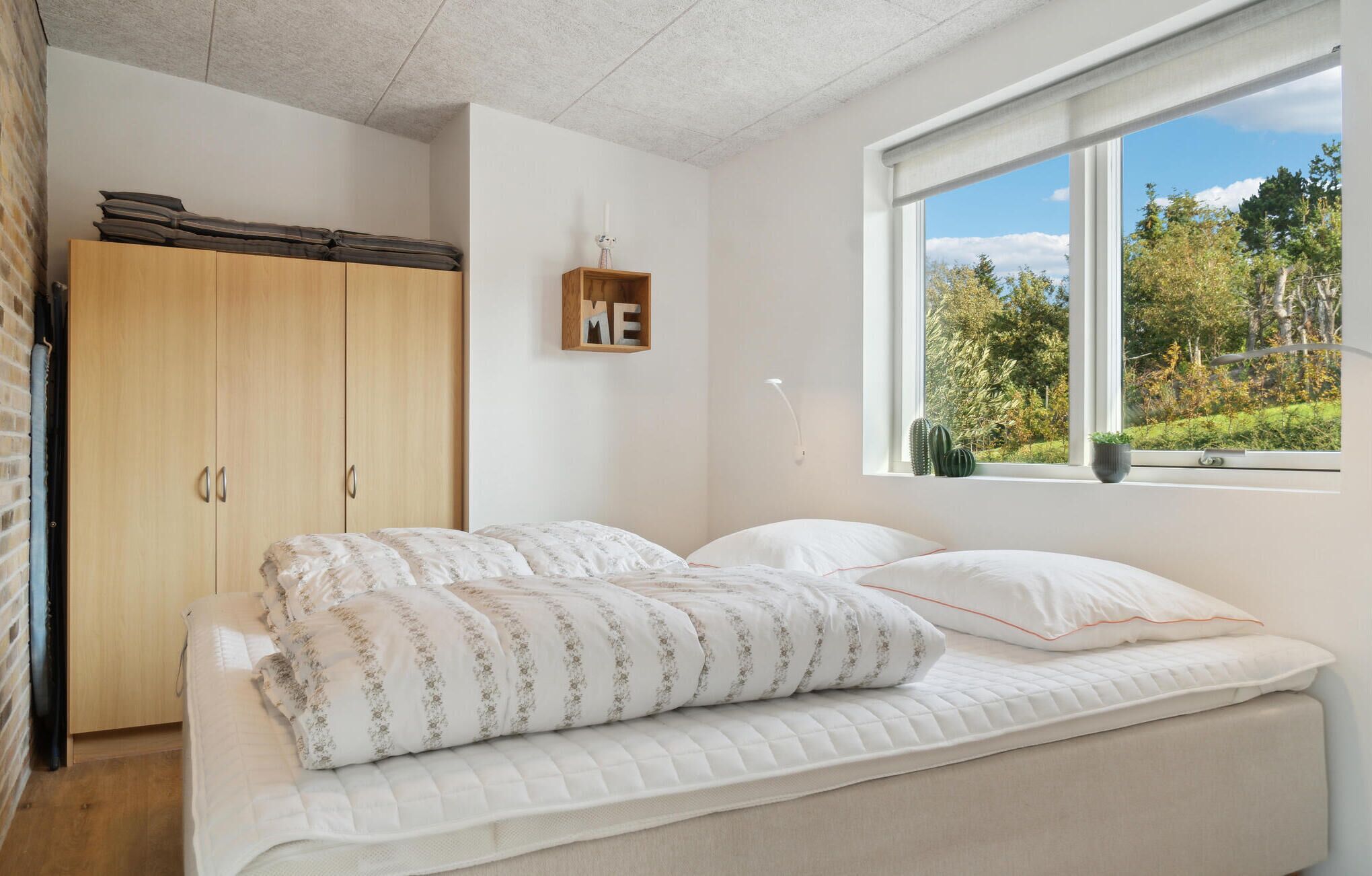 Une chambre avec un lit, une armoire, une fenêtre donnant sur des arbres et une étagère fixée au mur sur laquelle se trouve une bougie.