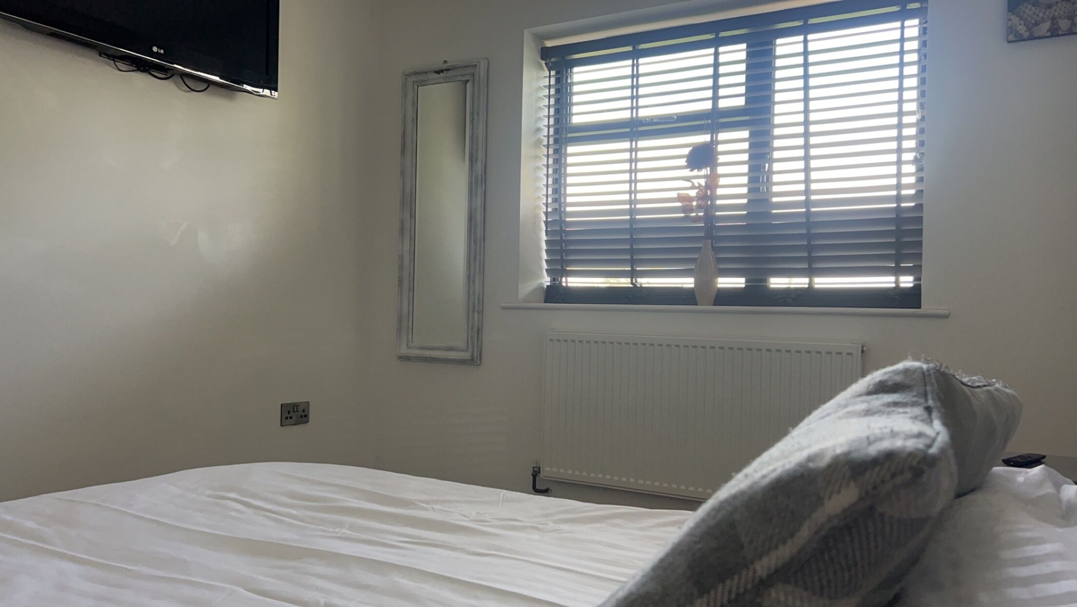 A bedroom with a bed, a window with blinds, a radiator, and a wall-mounted TV.
