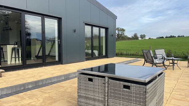 A modern house with large sliding glass doors, a patio area with outdoor furniture, and a view of a green field. - Beautiful 3-bed Barn in Uttoxeter (Uttoxeter)