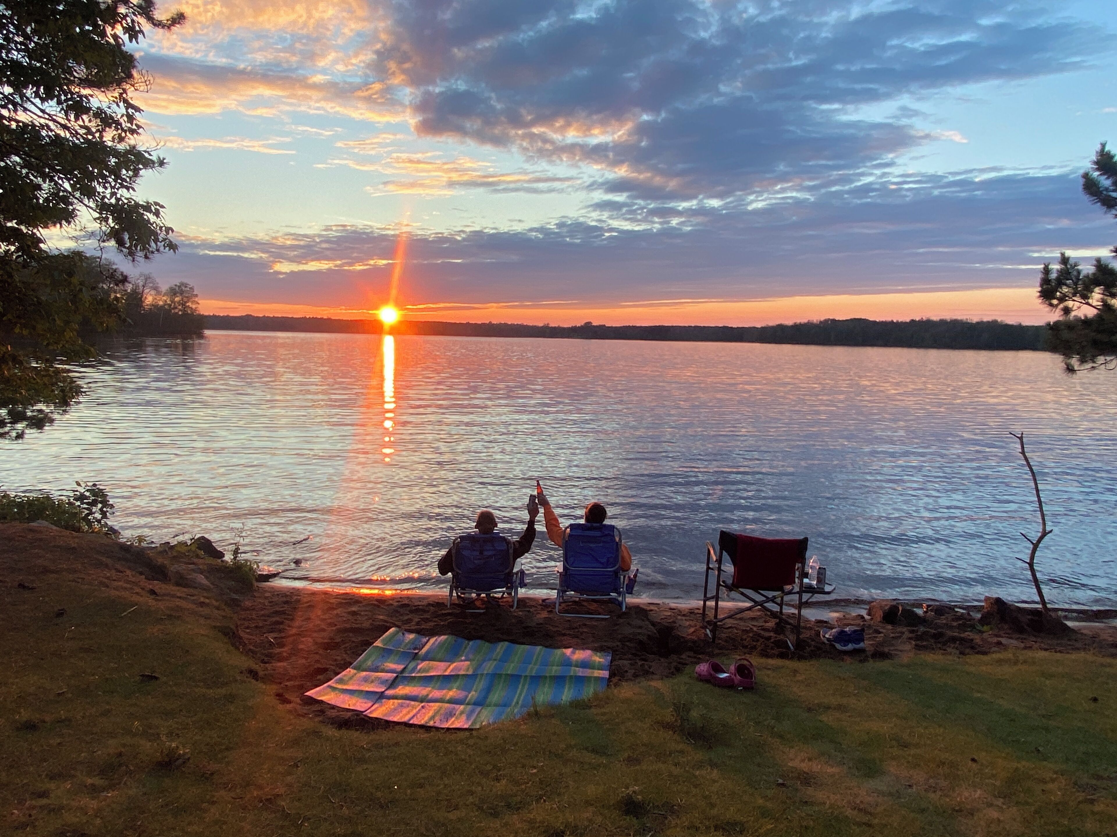 A sunset view over a lake with two people sitting on chairs facing the water, a blanket on the ground, and a pair of shoes nearby.