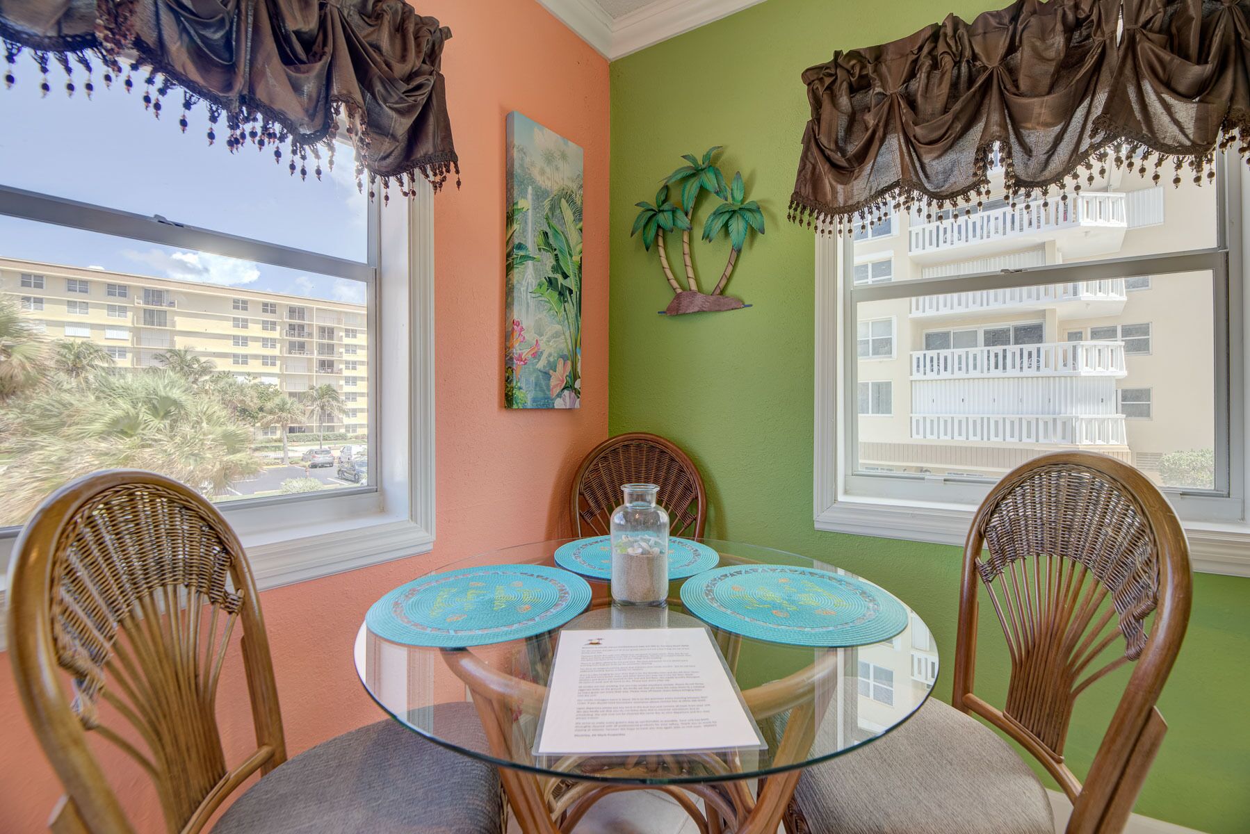 A room with a glass table, wicker chairs, a painting, and a view of a building.