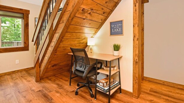 Una escalera de madera conduce a un dormitorio tipo loft con un escritorio y una silla, una ventana con vistas a la vegetación, y un cartel en la pared que dice 'life is better at the lake'.