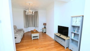 Una sala de estar con sofá, mesa de centro con fruta, televisor sobre un mueble, lámpara y reloj en la pared.