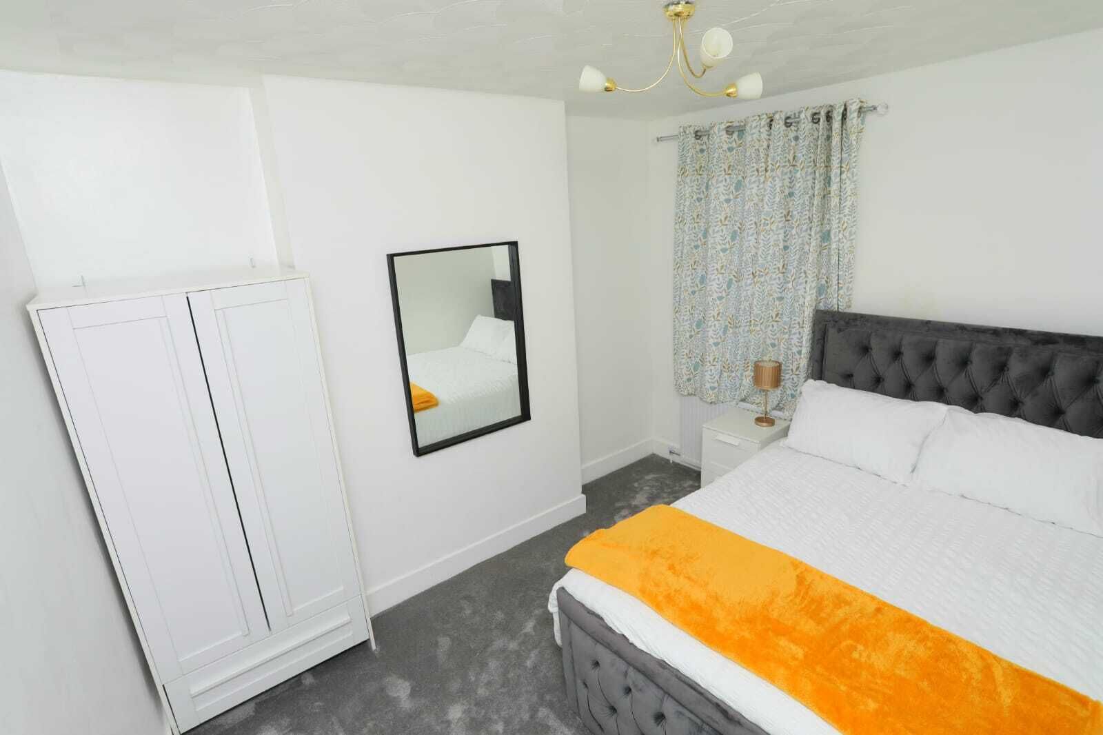 Un dormitorio con cama, armario, espejo, mesita de noche y ventana con cortinas.