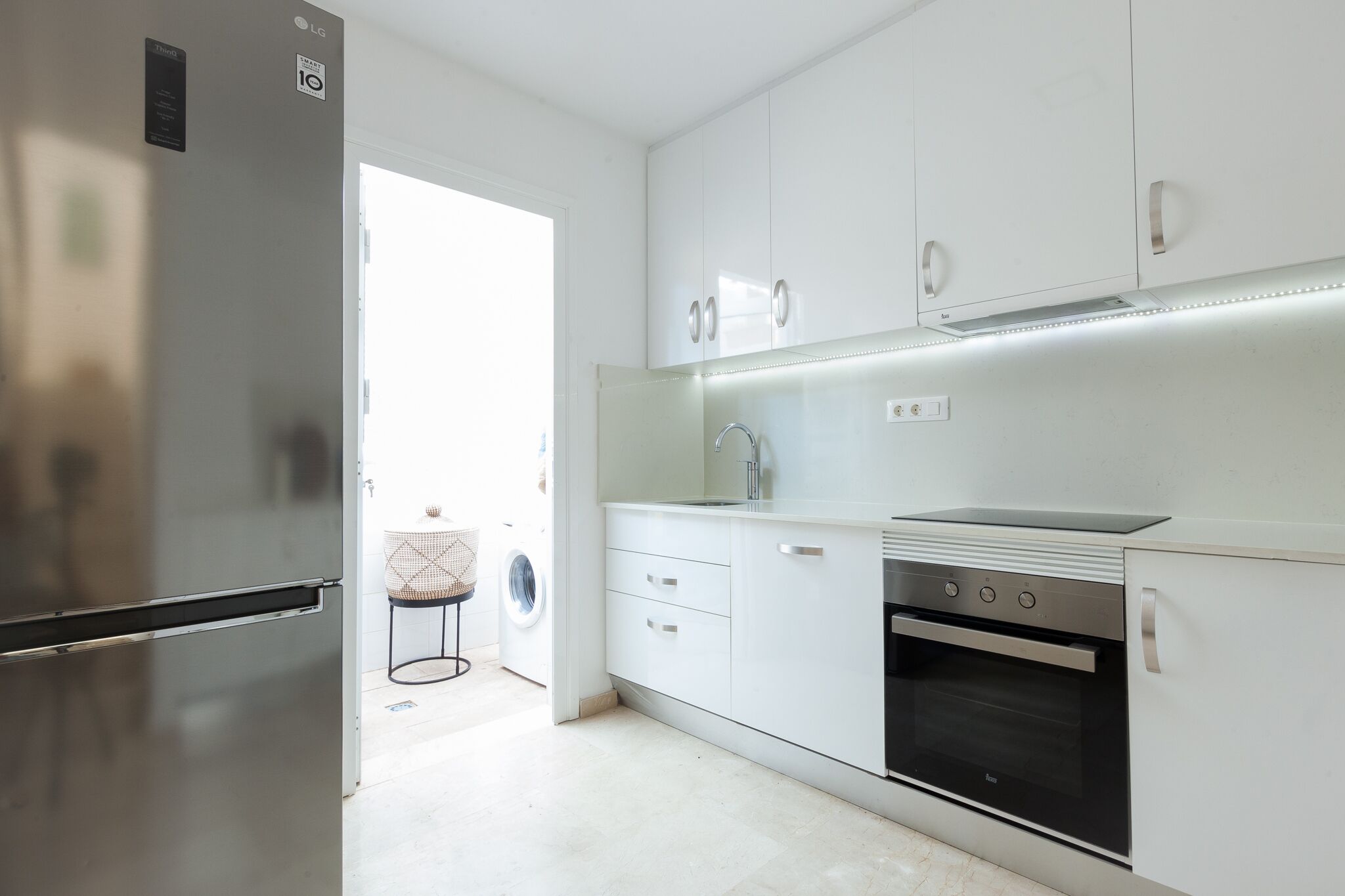 Ett modernt kök med vita skåp, ett kylskåp i rostfritt stål, en inbyggd ugn och en tvättmaskin i det intilliggande rummet.