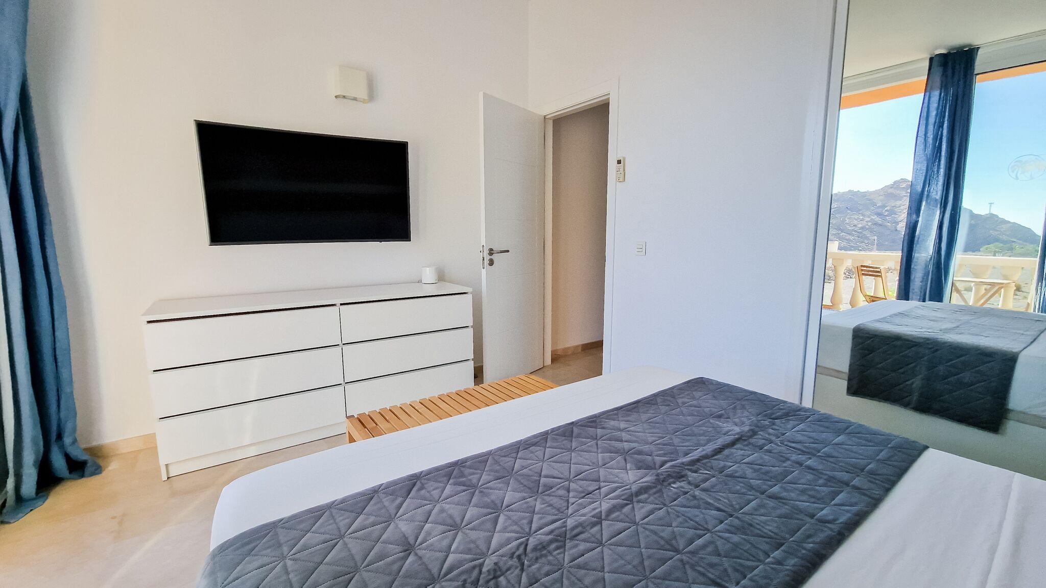 A bedroom with a bed, a TV mounted on the wall, a white dresser, and a balcony with a table and chairs.