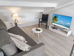Een moderne woonkamer met een grijze bank, een klein rond tafeltje met een decoratief object, een zwarte stoel en een flatscreen televisie waarop een strandscène te zien is.