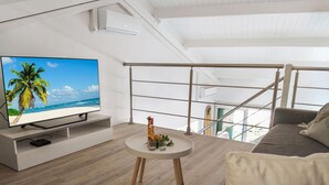 Un salon moderne doté d’un téléviseur à écran plat diffusant une scène de plage, d’une table ronde avec des boissons et des en-cas, d’un canapé et d’un escalier avec une rampe en verre.