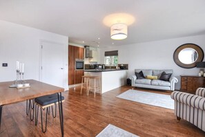 Una cocina moderna con una isla central, un comedor con mesa de madera y taburetes de bar, un sofá gris y un sillón a rayas.