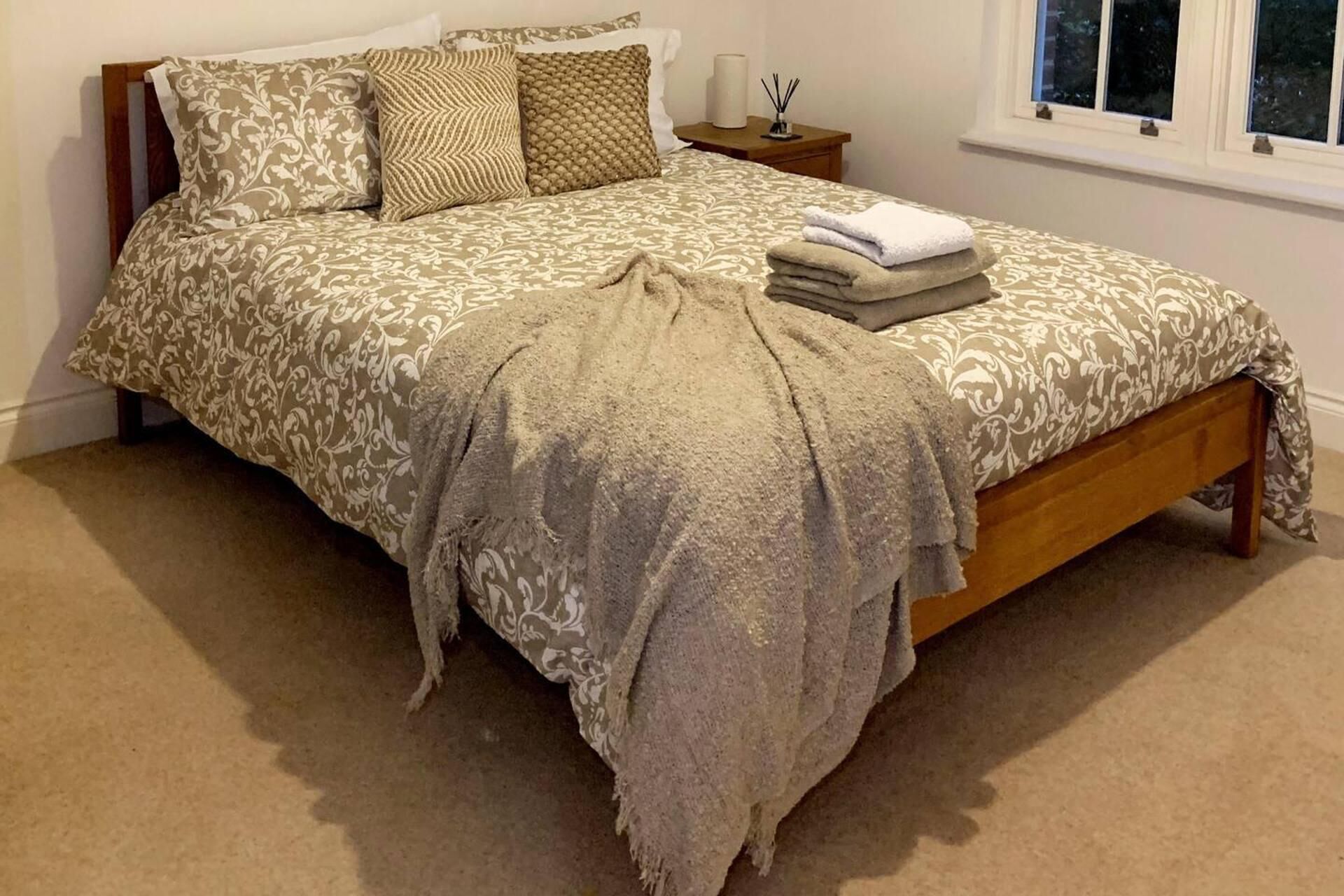 A neatly made bed with a patterned bedspread and pillows, a bedside table with a lamp and a small vase, and a window with a view of trees.
