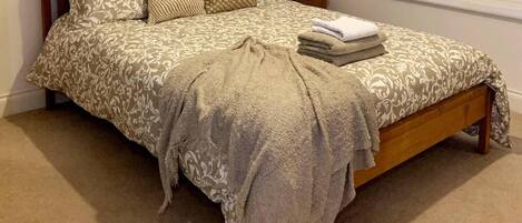 A neatly made bed with a patterned bedspread and pillows, a bedside table with a lamp and a small vase, and a window with a view of trees.