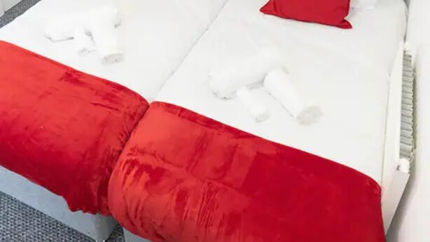 Quarto de hotel com duas camas, cada uma com um edredom vermelho e branco, e um cobertor vermelho no chão.