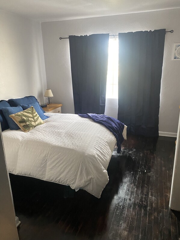 Ein Schlafzimmer mit einem Bett, dunklen Vorhängen, einem Nachttisch mit Lampe und Holzboden.