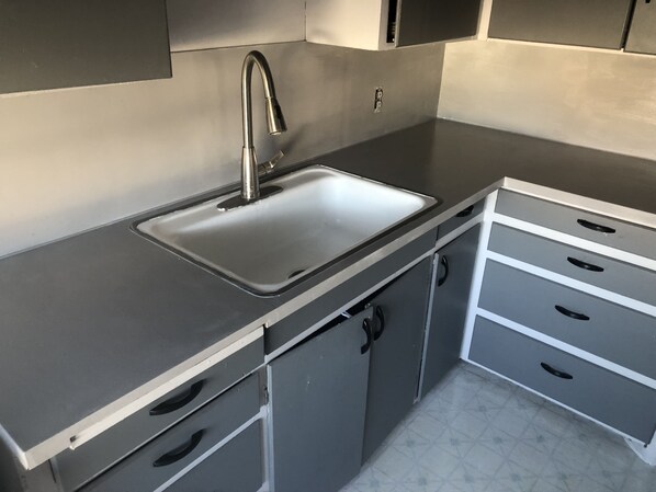 Eine Küche mit Spüle aus Edelstahl, einem Wasserhahn und einer Arbeitsplatte. Darunter befinden sich drei Schubladen und ein Schrank.