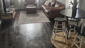A living room with a brown leather sofa, a small round table with white chairs, a ceiling fan, and a window with patterned curtains.