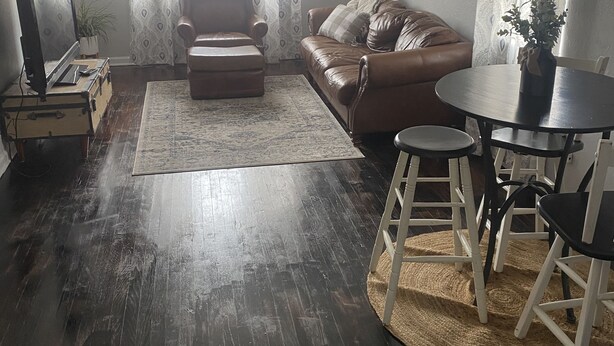 Un salon avec un canapé en cuir marron, une petite table ronde entourée de chaises blanches, un ventilateur de plafond et une fenêtre ornée de rideaux à motifs.