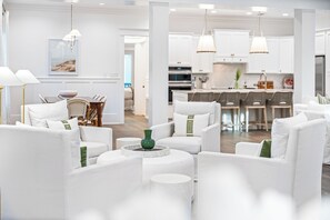 A modern living room with white furniture, a dining area, and a kitchen with white cabinets and pendant lighting.