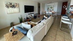 Un salon moderne avec un canapé blanc, un téléviseur à écran plat, un ventilateur de plafond et un coin repas avec un bar.